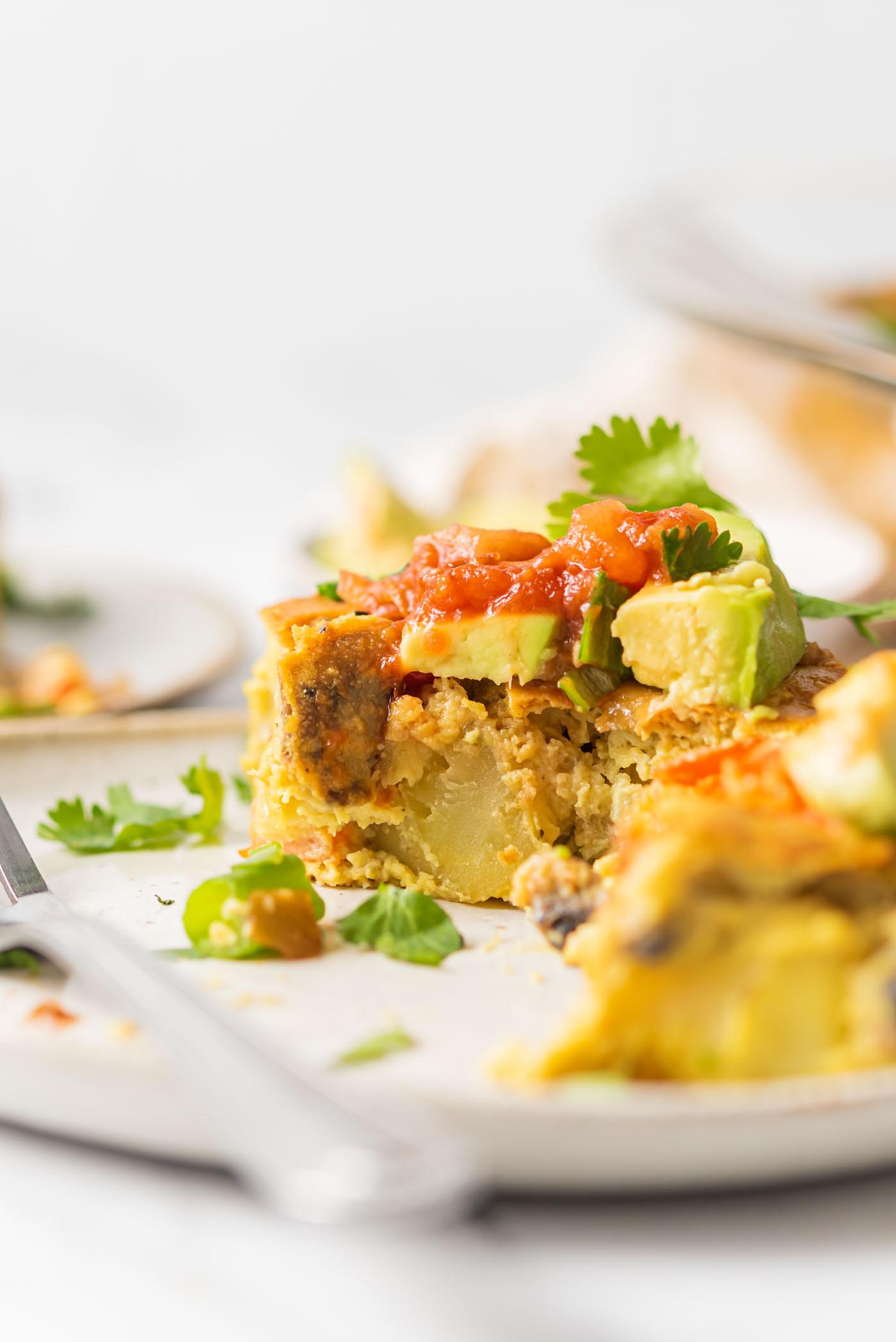 Sny van vegan eierlose kasserol met stukkies aartappel en groente daarin, bedek met avokado en salsa.