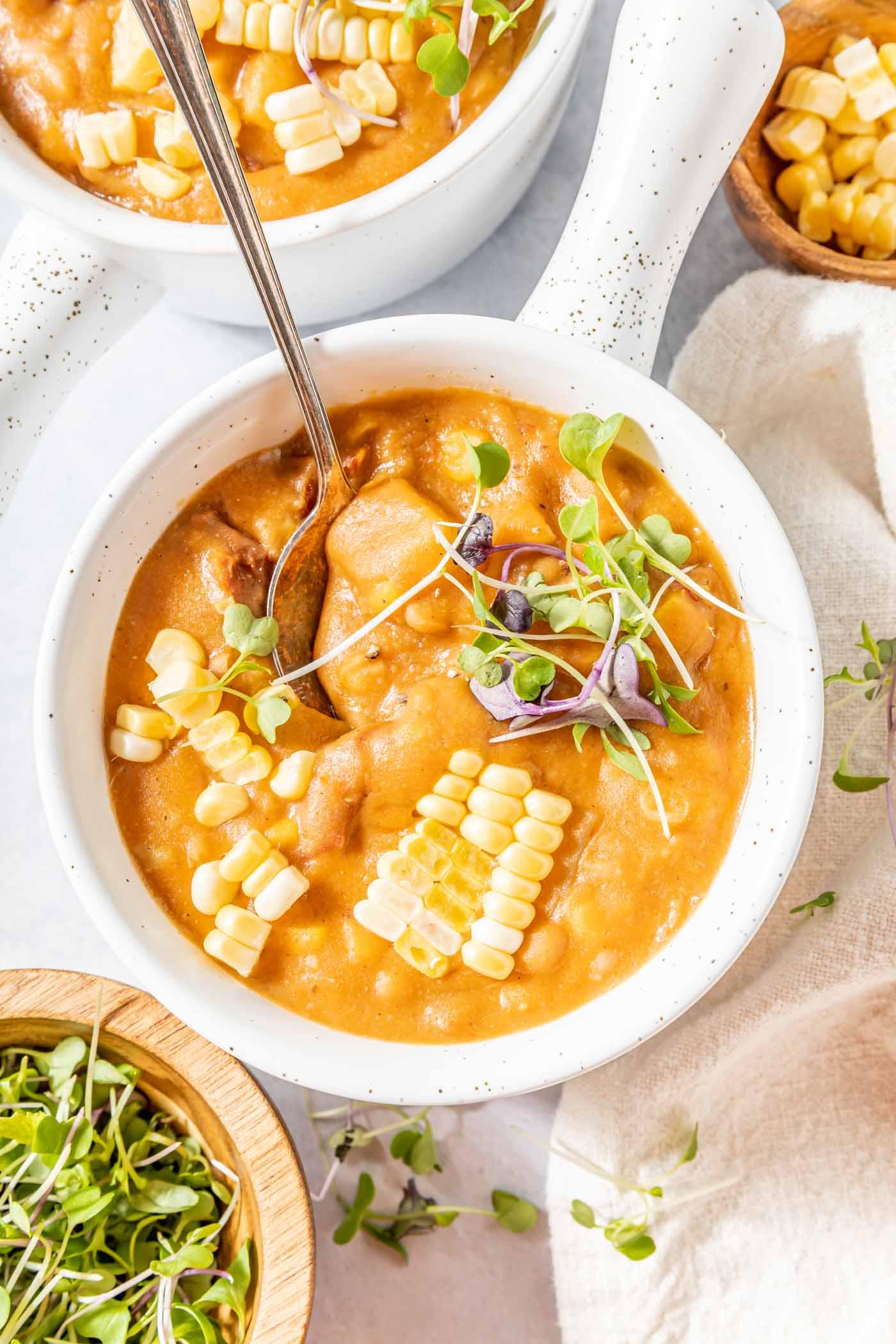 Vegan Somer Mielieroomop 2 Oorsig van 'n bak somerkoringchowder sop bedek met mikro-groente en vars mieliepitte. 'n Lepel rus in die bak en twee addisionele bakke sop is net buite die raam.