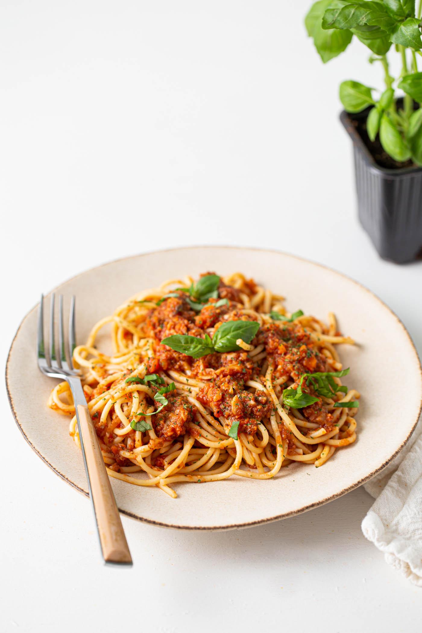 Bord vegan sampioen bolognese sous oor spaghetti met basiliekruid en parmesaan bo-op. 'n Gaffel rus op die bord en daar is 'n vars basiliekruidplant op die agtergrond.