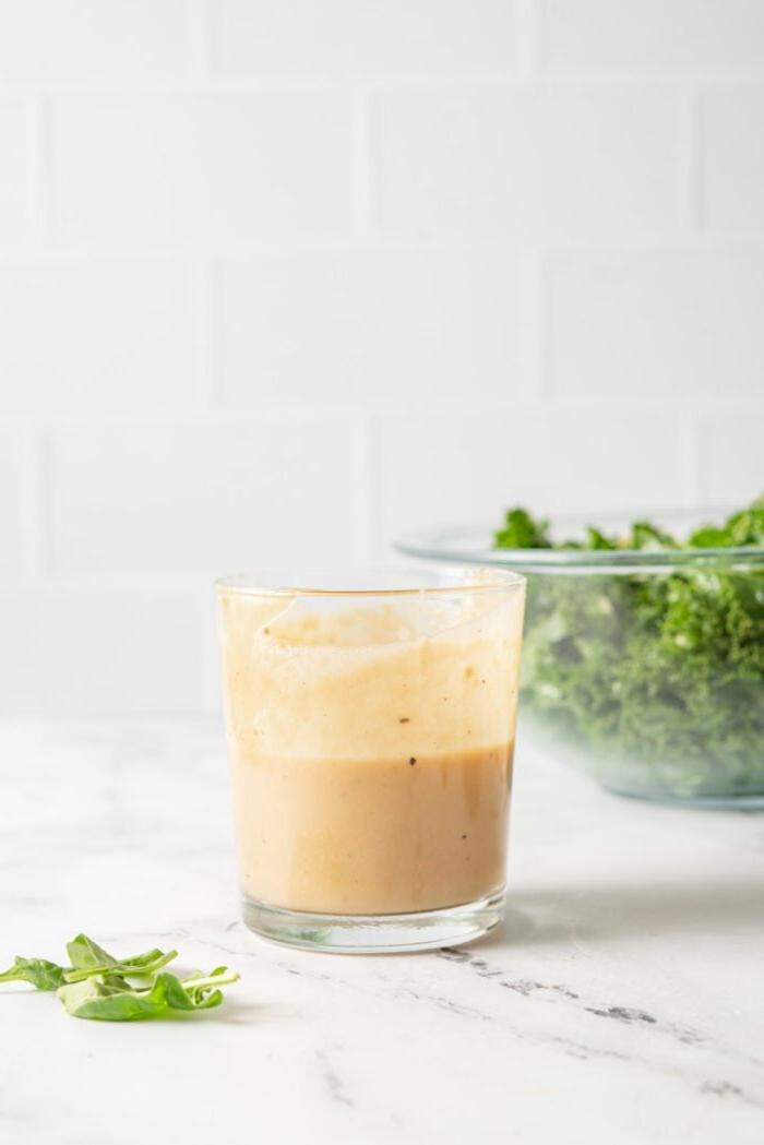 Fles romerige tahini-esdoorn-dijon-mosterdsous op 'n marmerblad met 'n bak gekapte boerenkool in die agtergrond.