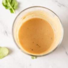 Oorhoofse aansig in 'n fles romerige tahini-dijon-esdoorn-slaaisous met 'n paar stukkies spinasie rondom dit.
