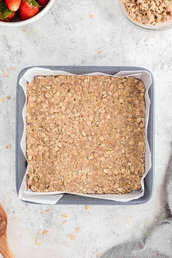 Vegan Aarbei Krummel Repies 5 ’n Laag hawermoutkors in ’n vierkantige bakpan gedruk wat met perkamentpapier uitgevoer is.