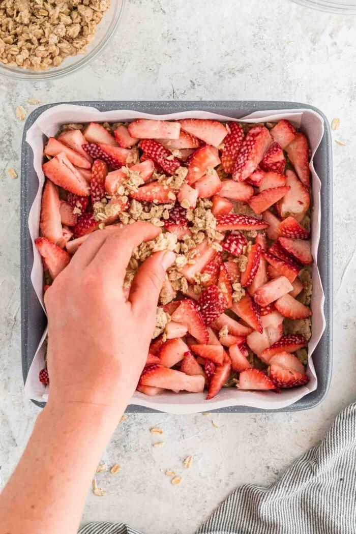 Vegan Aarbei Krummel Repies 7 ’n Hand wat ’n hawermoutkrummelmengsel oor gesnyde aarbeie in ’n vierkantige bakpan strooi wat met perkamentpapier uitgevoer is.