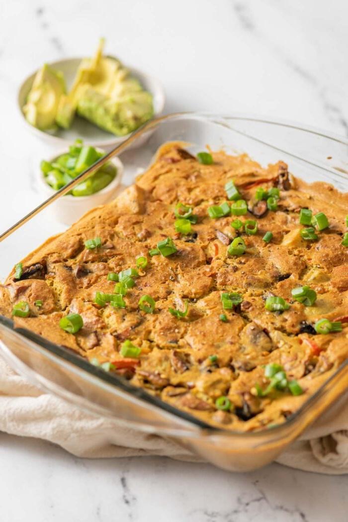 Gebakte vegan aartappel eierkasserol in 'n glasbakskottel. Klein bakkie gesnyde avokado is in die agtergrond.