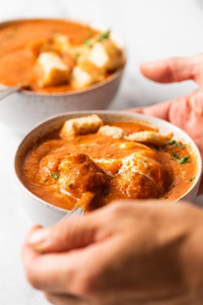 Geroosterde Rooipeper Tamatie Sop 8 Close up of hand taking a spoonful of roasted red pepper tomato soup from a bowl with a spoon. Another bowl of soup appears in background.