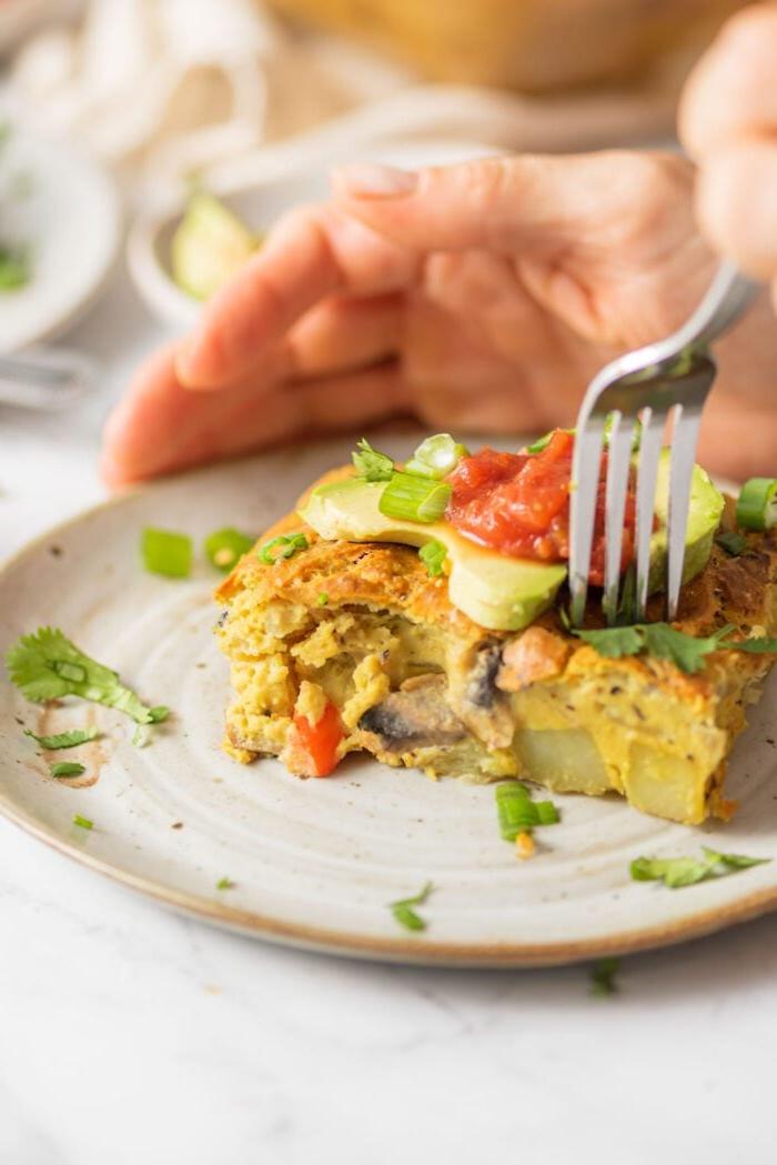 Sny van vegan ontbyt kasserol met aartappel en groente bo-op met avokado en salsa. Hand is besig om 'n vurk te gebruik om 'n sny daarvan te neem.