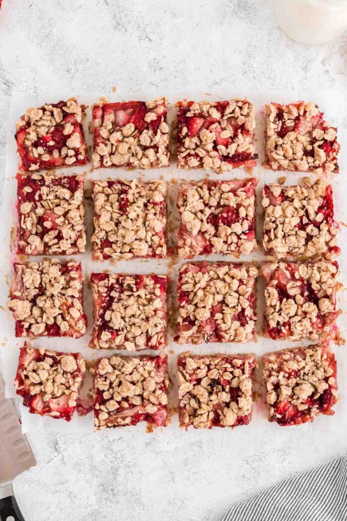 Vegan Aarbei Krummel Repies 9 Aarbei-hawermoutstafies in 16 egalige blokkies op 'n stuk perkamentpapier gesny. 'n Mes rus langs die stafies.