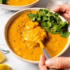 Suurlemoenlensiesop 10 Hand gebruik 'n lepel om 'n lepelvol suurlemoen lensiesop, bedek met vars koljander, uit 'n bak te lig. Nog 'n bak sop is in die agtergrond.