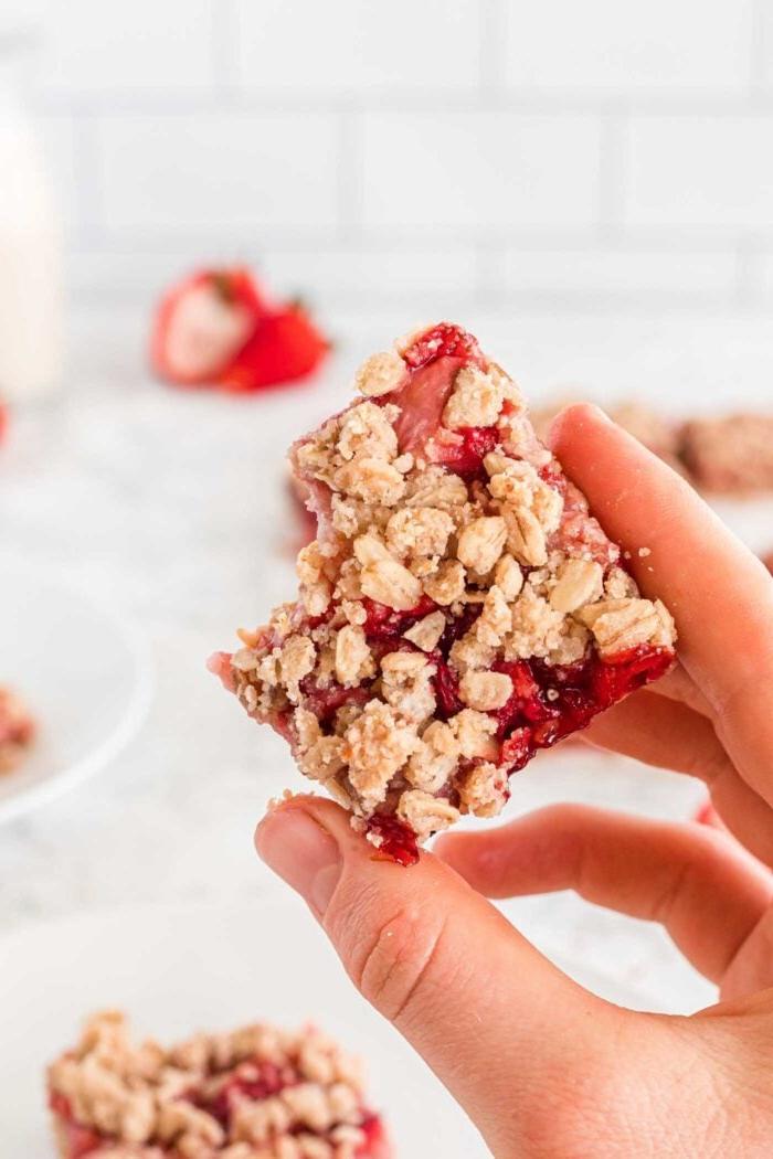 Vegan Aarbei Krummel Repies 10 ’n Hand wat ’n aarbeikrummelstafie vashou met ’n hap daaruit. Daar is ’n paar vars aarbeie op die agtergrond en ’n bord met meer stafies daarop.