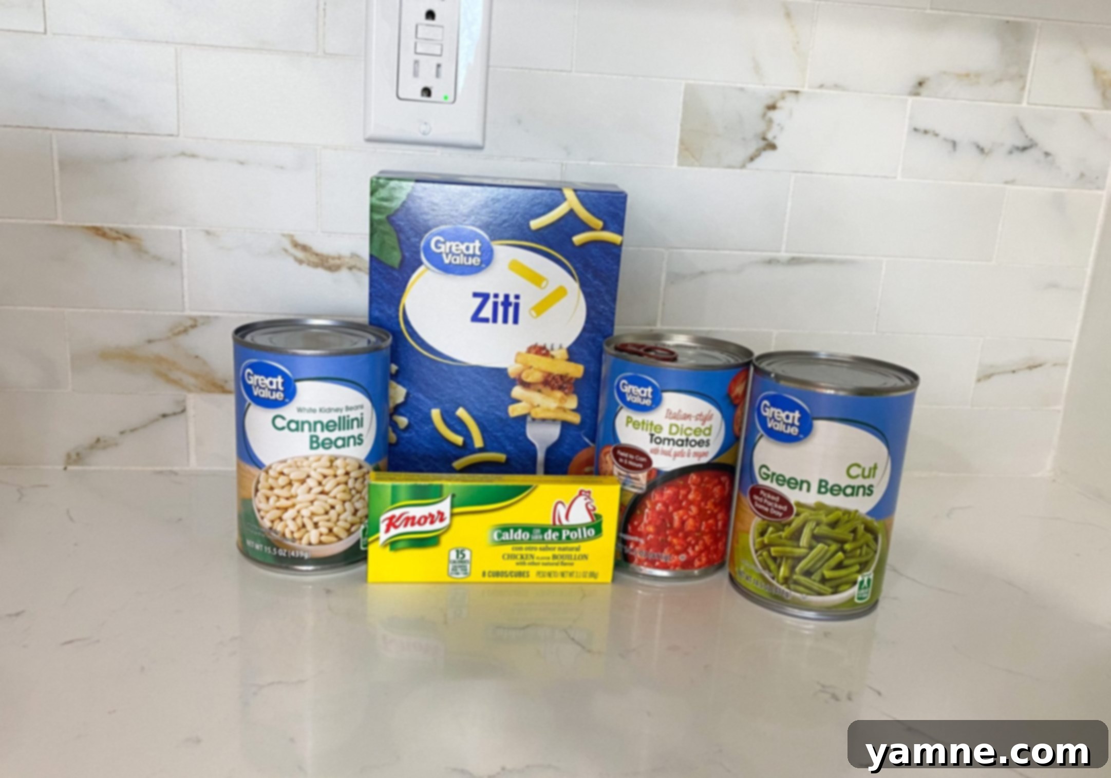 A selection of simple ingredients laid out for vegetable noodle soup, including dry pasta, canned cannellini beans, canned diced tomatoes, and canned green beans.