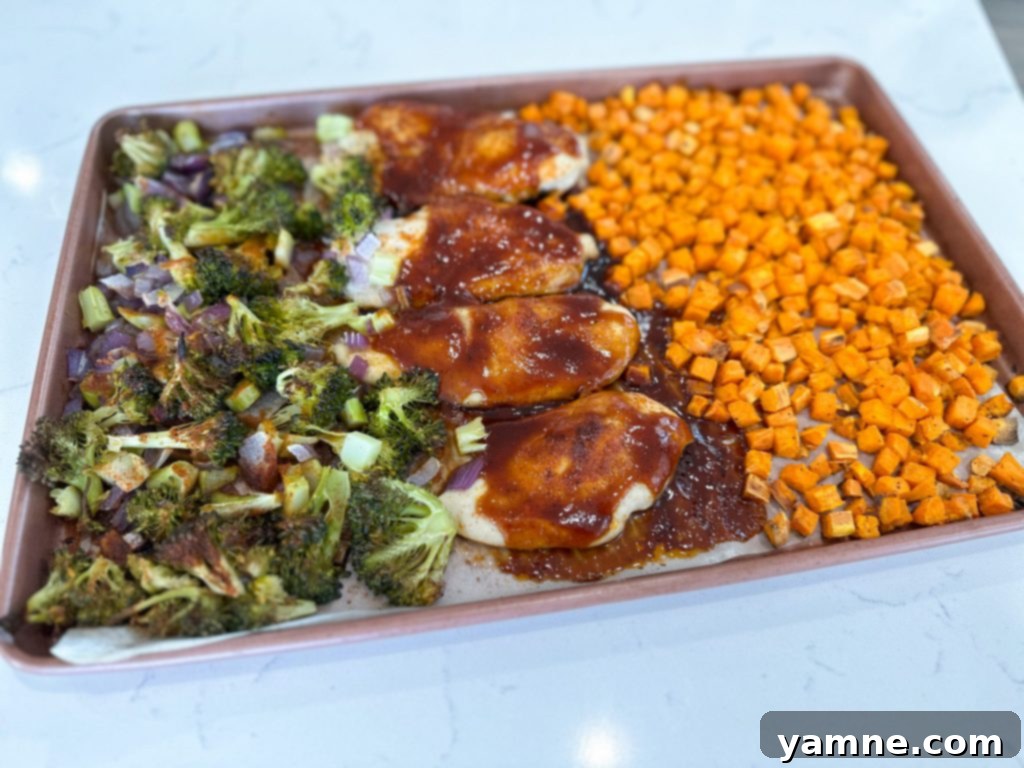 Close-up view of raw chicken breasts seasoned with a spice rub, alongside diced sweet potatoes, broccoli, and onions on a parchment-lined sheet pan.
