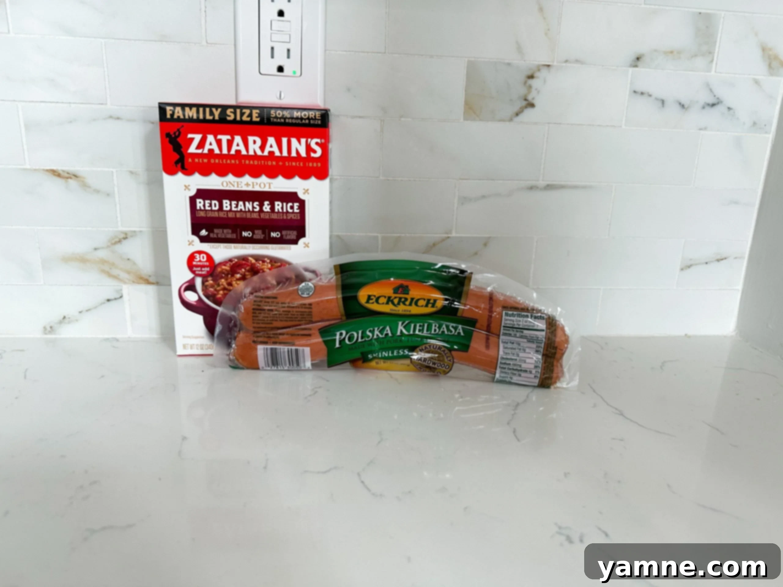 A flat lay of the four simple ingredients for red beans and rice: a pack of Kielbasa sausage, a box of Zatarain's Red Beans and Rice mix, a bottle of olive oil, and a measuring cup of water.