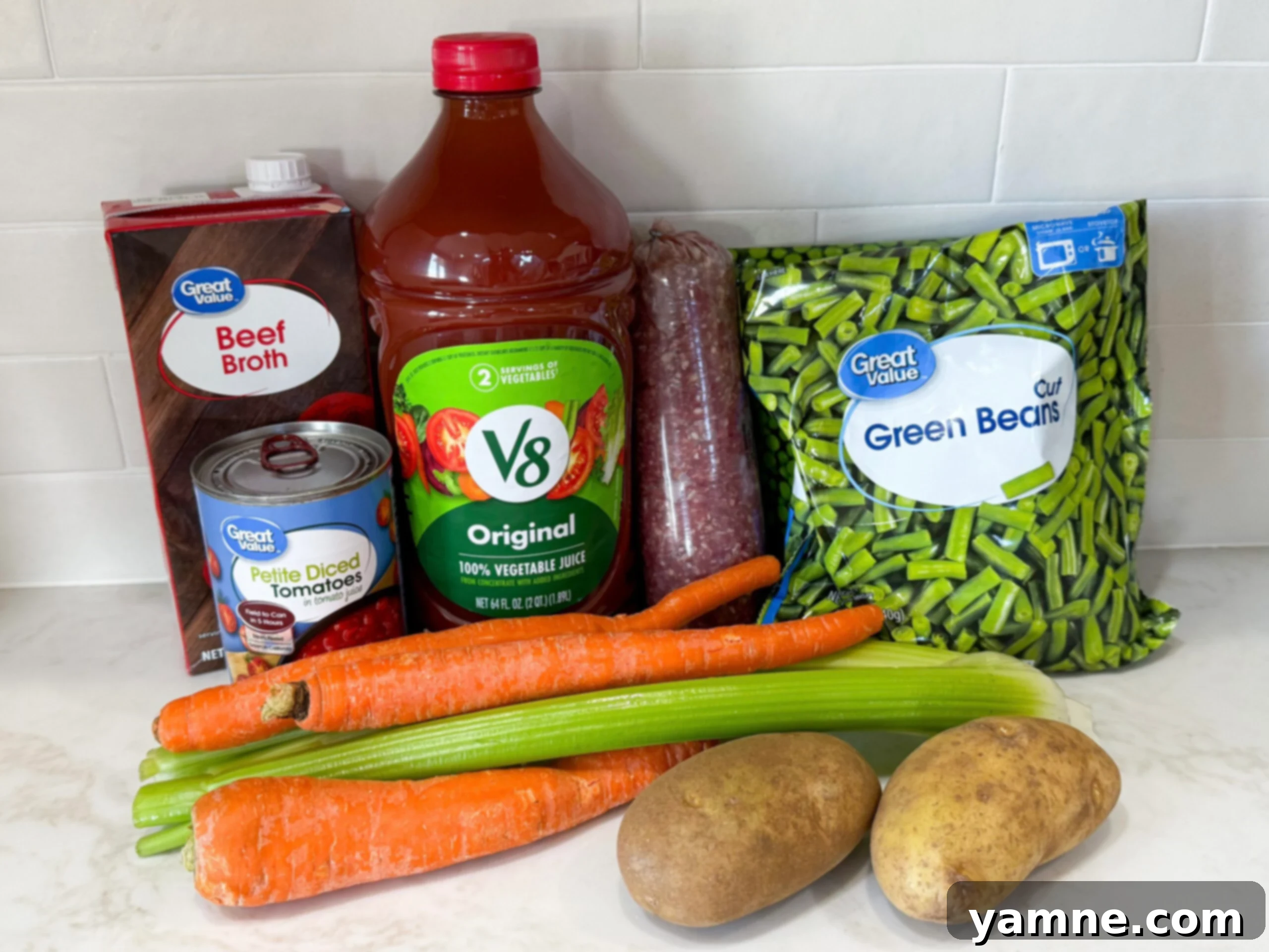 Fresh ingredients meticulously laid out for a comforting ground beef and vegetable soup, including ground beef, diced potatoes, sliced carrots, celery, onion, V-8 juice, and various spices.