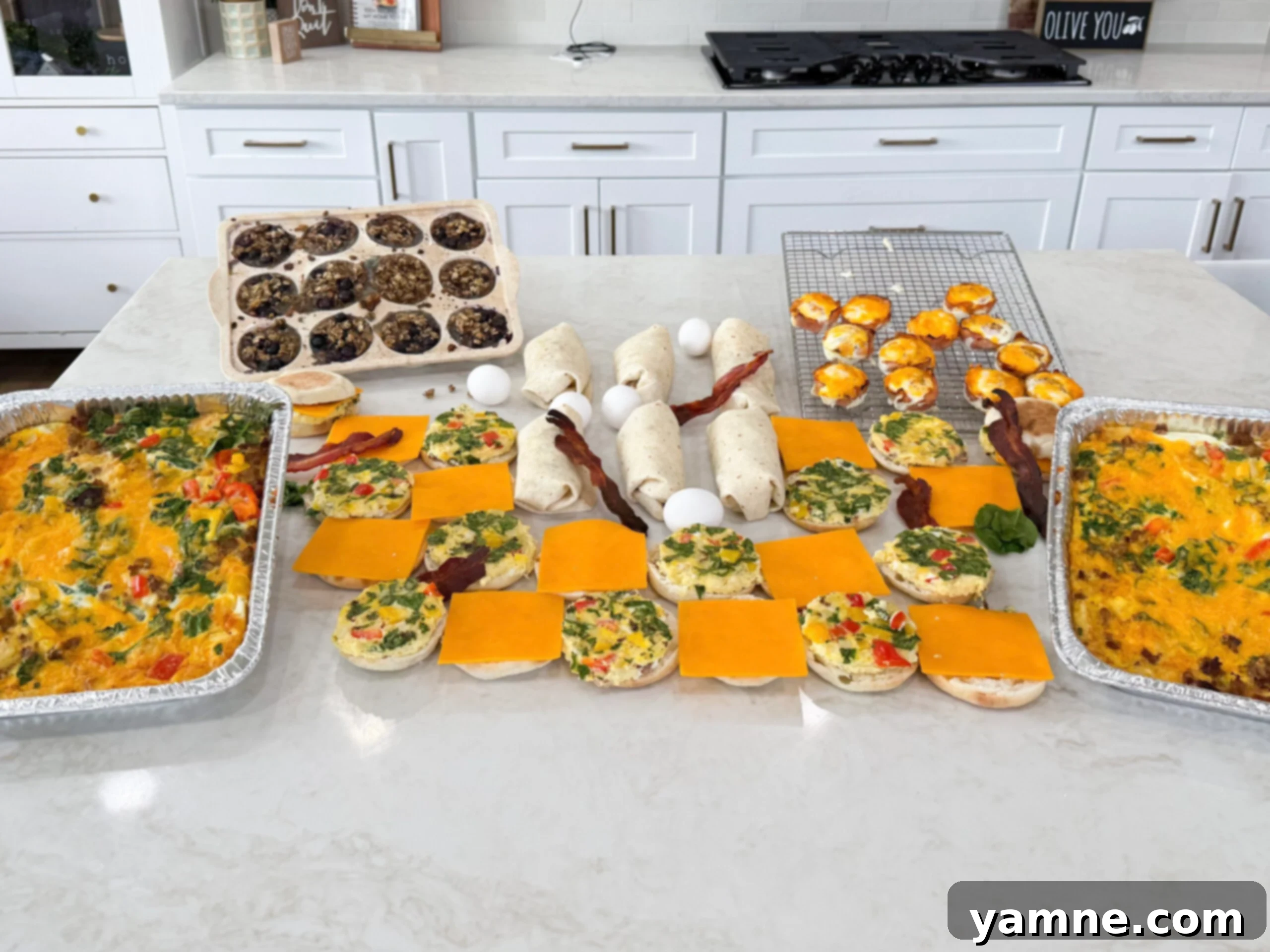 Completed breakfast meal prep including two breakfast casseroles, baked blueberry oatmeal cups in a muffin tin, ham and egg bites on a cooling rack, breakfast burritos, and breakfast sandwiches on a white countertop.