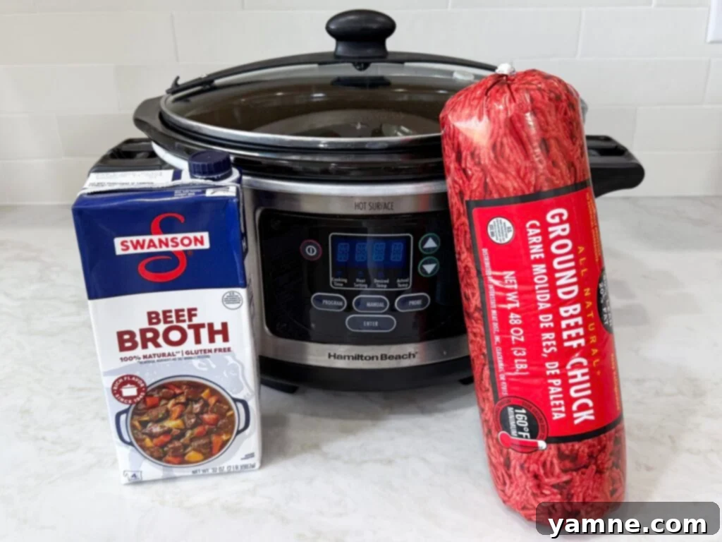 Large slow cooker on a white counter with a package of ground beef and a carton of beef broth, ready for preparation of the slow cooker ground beef recipe.