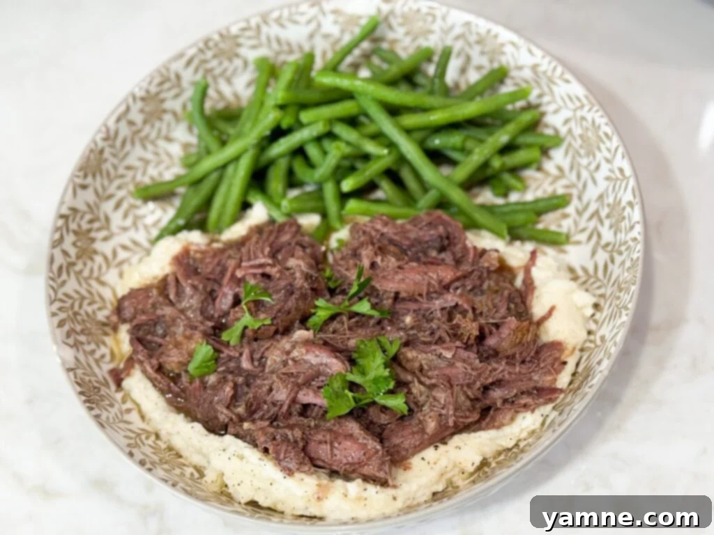 A serving of tender shredded beef over creamy mashed potatoes with a side of green beans, presented on a stylish grey and white plate.