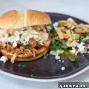 A dark plate holds slow cooker Hawaiian barbecue chicken sandwiches with a side salad on a grey counter. The chicken is shredded and coated in a sweet and tangy sauce.