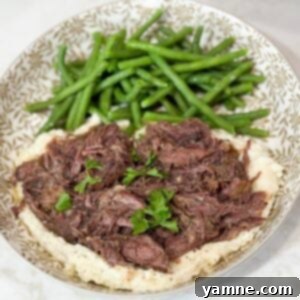 Shredded beef served over a bed of creamy mashed potatoes with vibrant green beans on a modern grey and white plate.