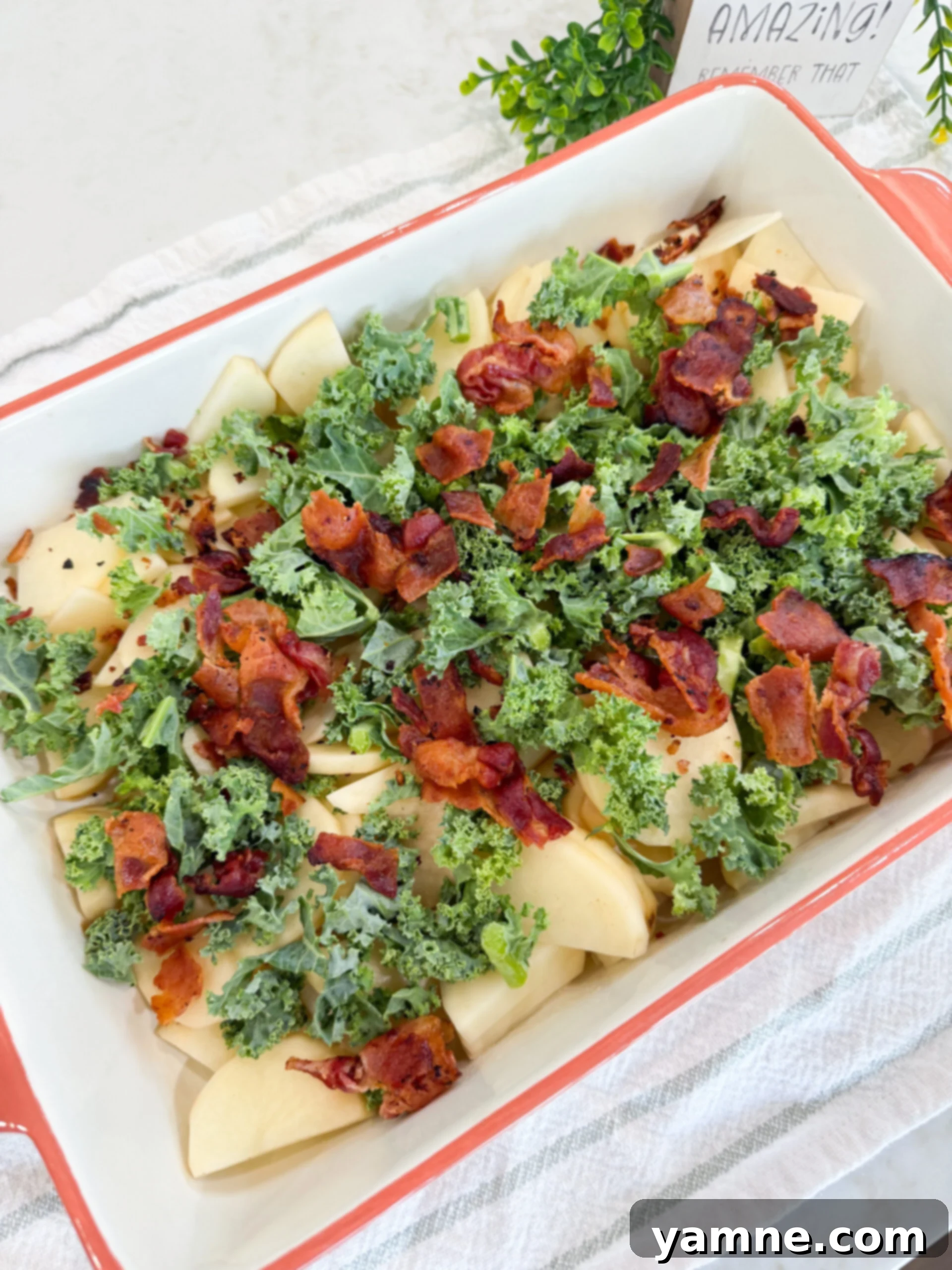 Creamy Zuppa Toscana Sausage Potato and Kale Bake 8 Chopped kale and crumbled crispy bacon layered over thinly sliced raw potatoes in a baking dish, ready for the sauce.