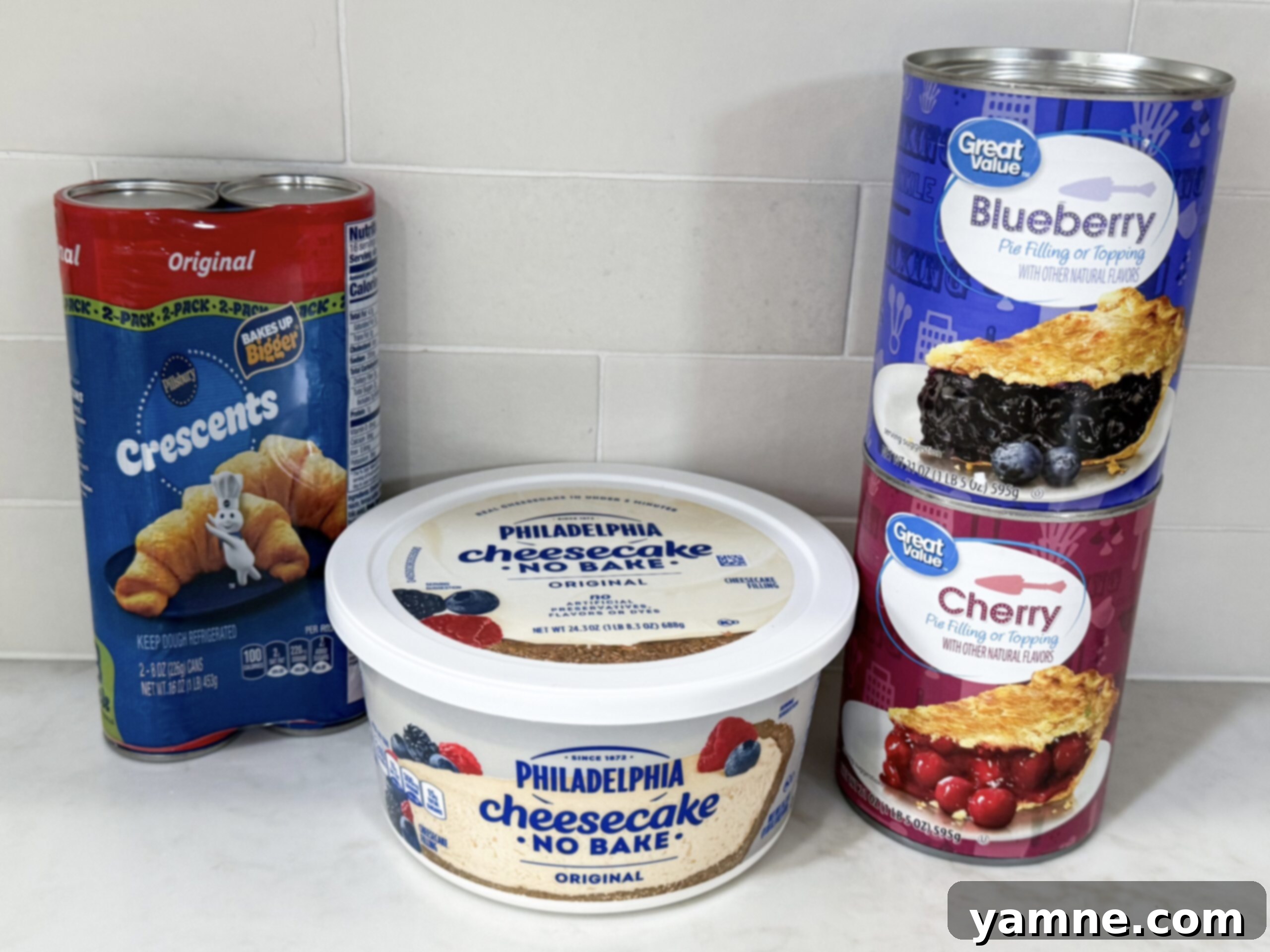 All the simple ingredients for mini cheesecake pies laid out on a kitchen counter, including crescent dough, cheesecake filling, and various fruit toppings.