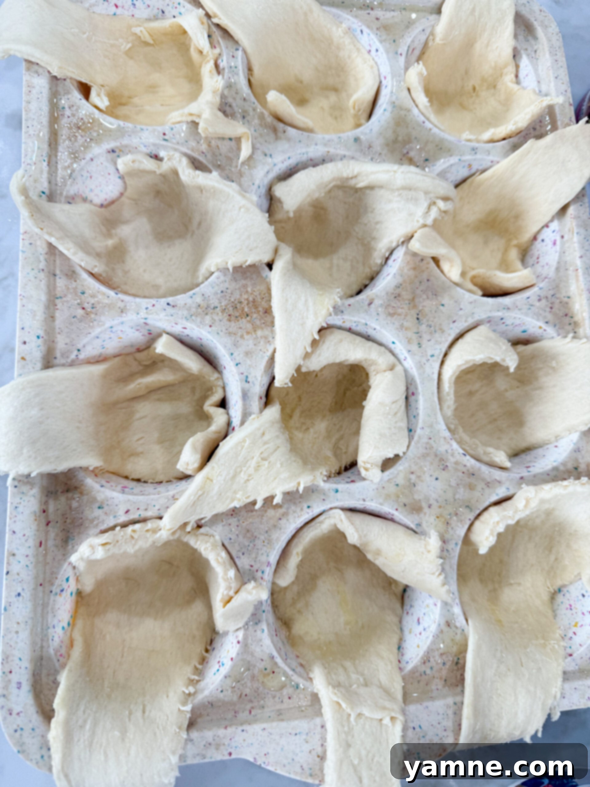 A hand gently pressing a crescent dough triangle into a muffin tin cup, forming the base for a mini cheesecake pie.