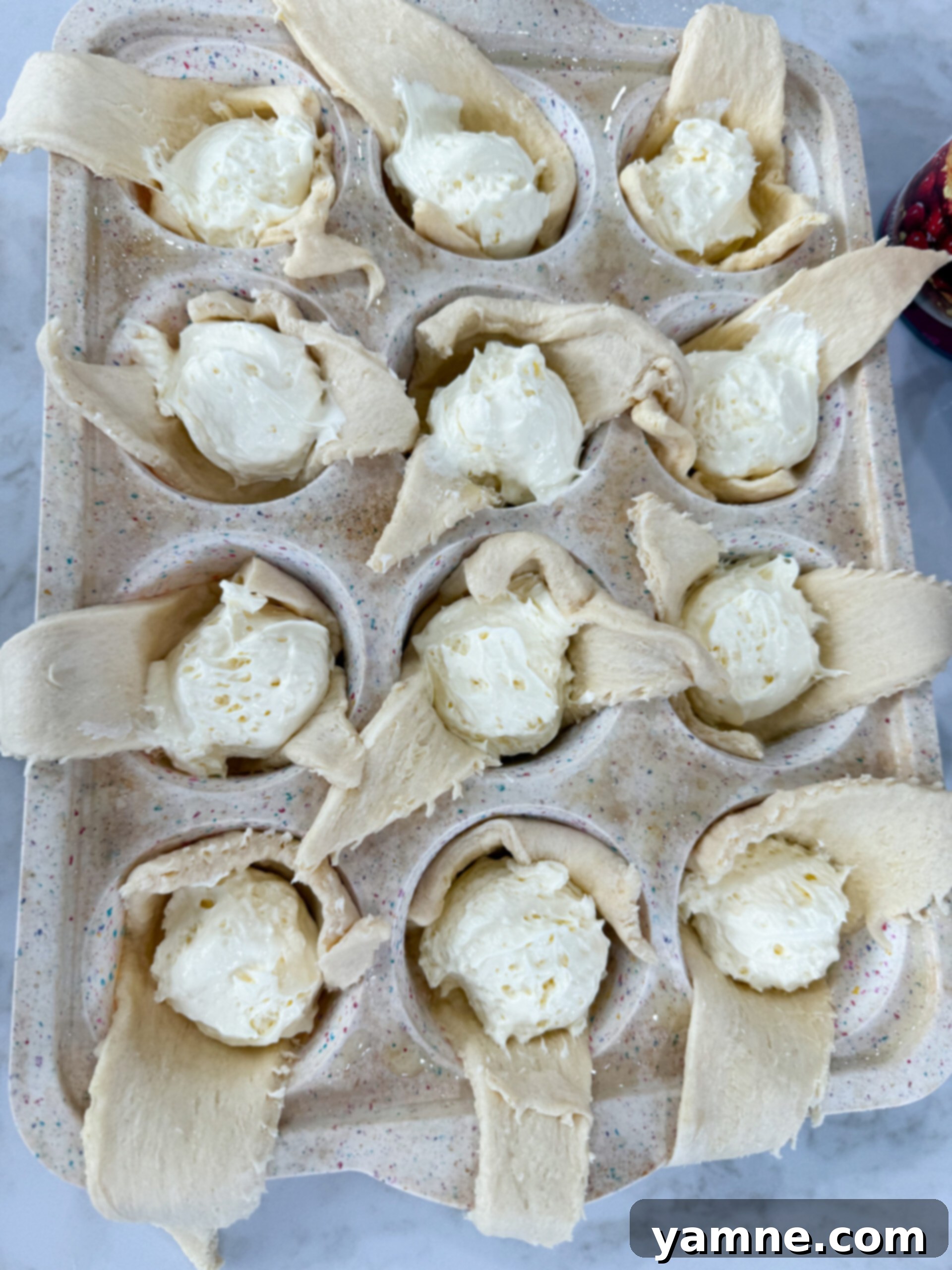Muffin cups filled with crescent dough, being spooned with creamy cheesecake filling.