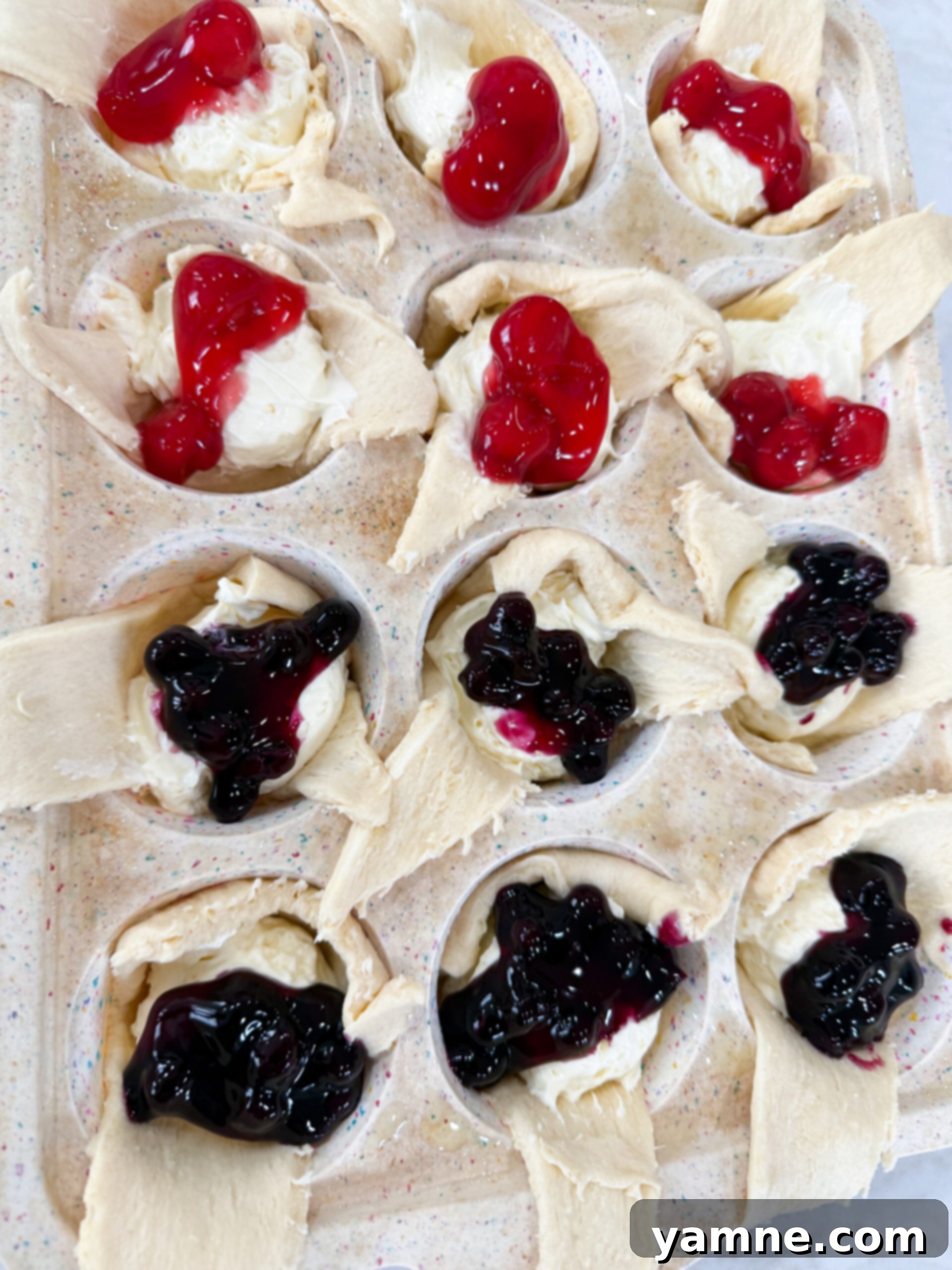 Mini cheesecake pies, partially baked, being topped with generous spoonfuls of blueberry and raspberry pie filling.
