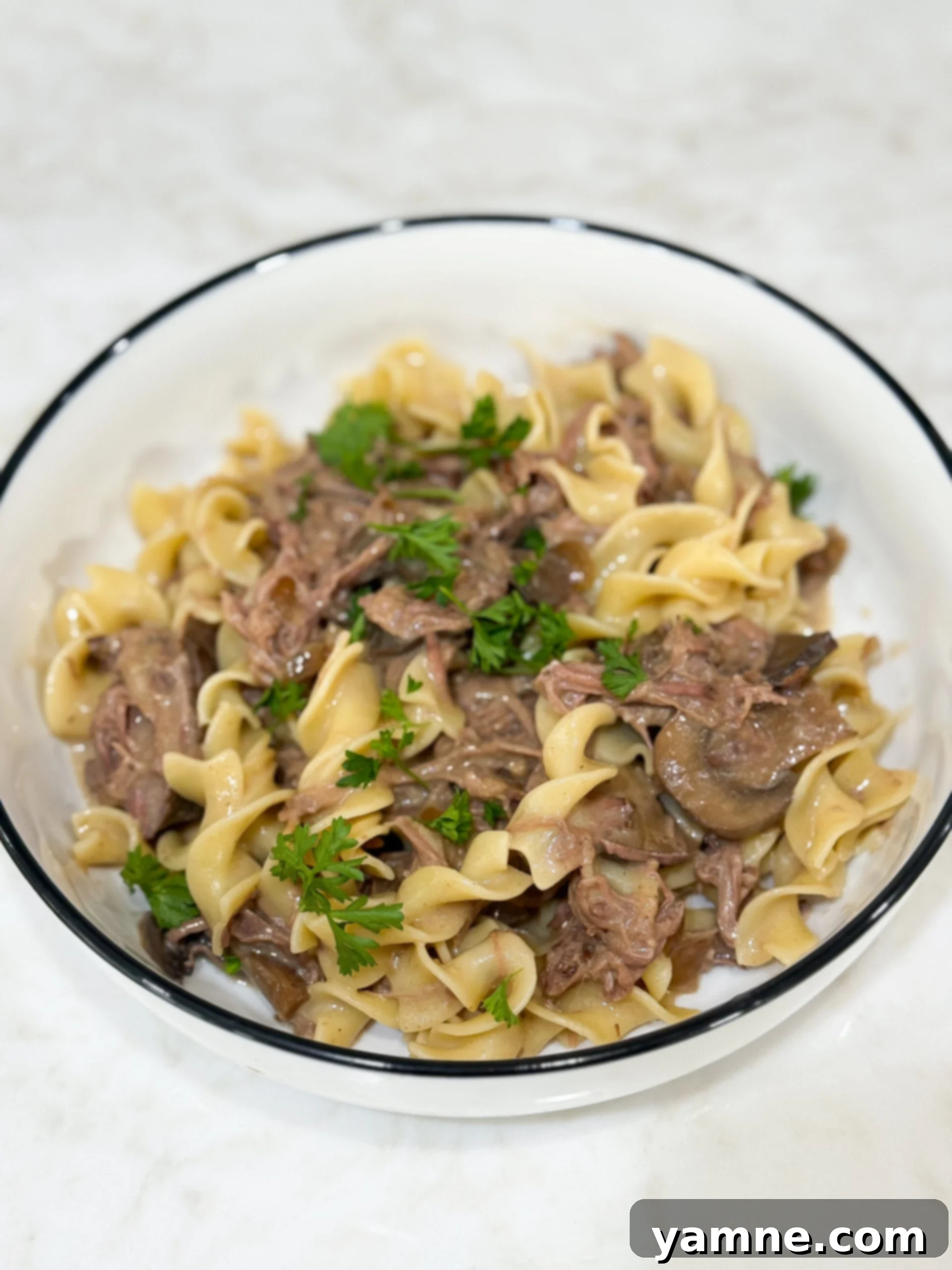 Easy Slow Cooker Beef Stroganoff 2 Slow cooker beef stroganoff served with noodles and fresh parsley garnish in a white bowl on a wooden surface.