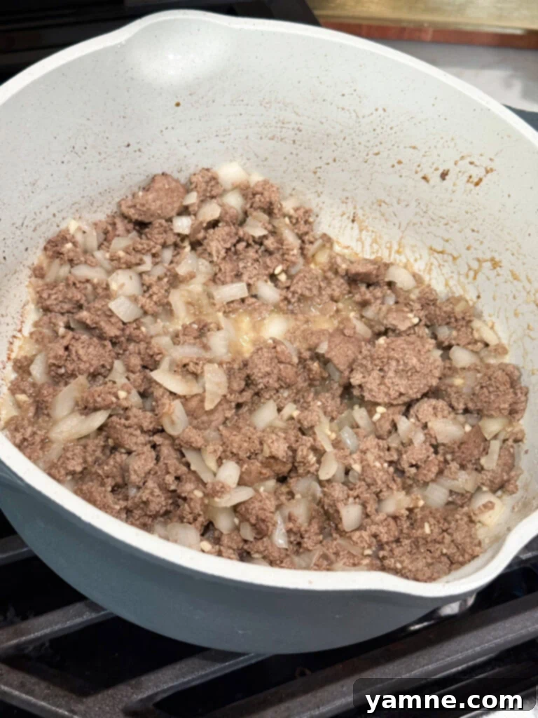 Ground beef and onions browning together in a large skillet on the stovetop, releasing their savory aromas.