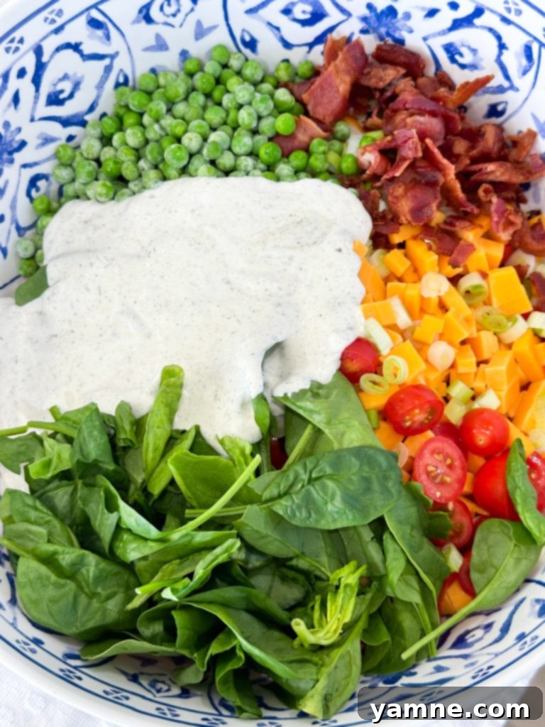 All the fresh ingredients, including cooled pasta, bacon, cheese, tomatoes, and spinach, being added to a large bowl with creamy ranch dressing.