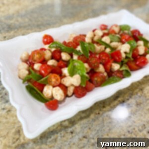 A vibrant Caprese salad featuring sliced tomatoes, fresh mozzarella pearls, and green basil leaves, drizzled with balsamic glaze and olive oil, served in a white bowl.